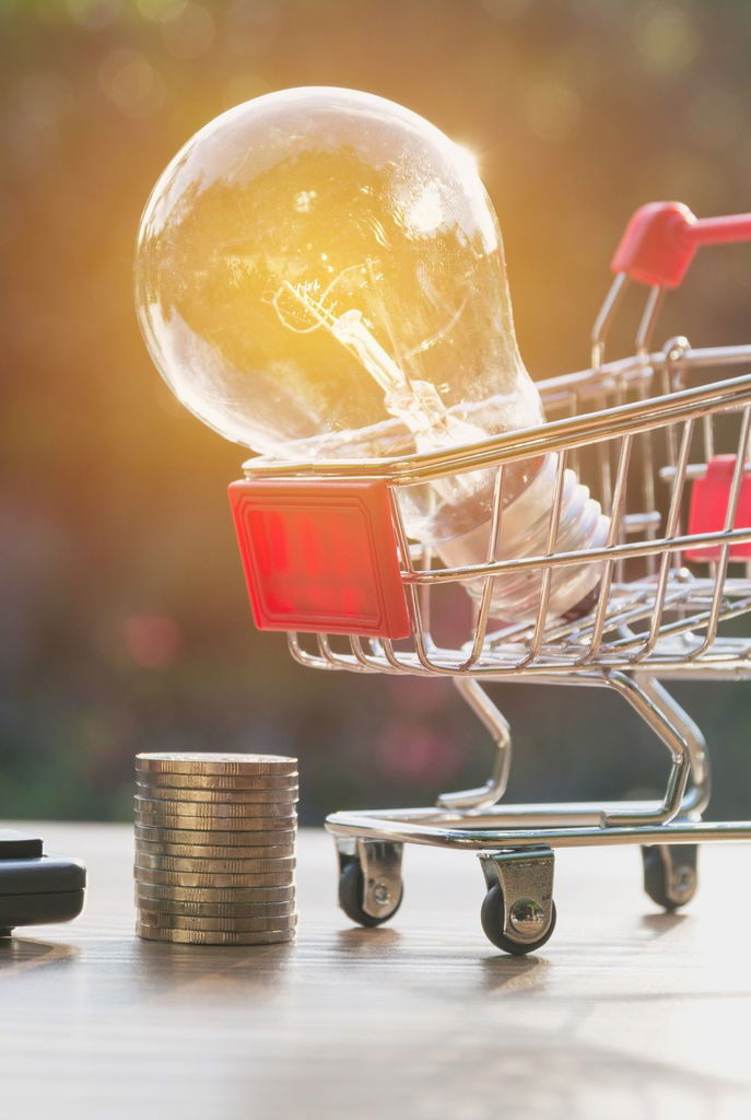 A shopping cart with lighted light bulb beside stack of coins and calculator